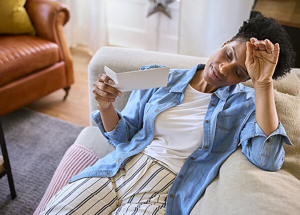Reife, Schwarze Frau in den Wechseljahren, die zu Hause auf dem Sofa sitzt und sich selbst mit einem Umschlag Luft zufächelt.
