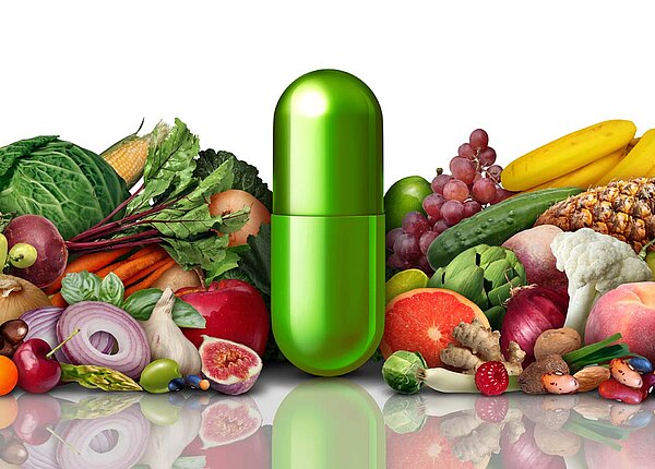 Grüne Medikamentenkapsel steht hochkant in der Mitte: Um die Kapsel herum liegt verschiedenes Obst und Gemüse.