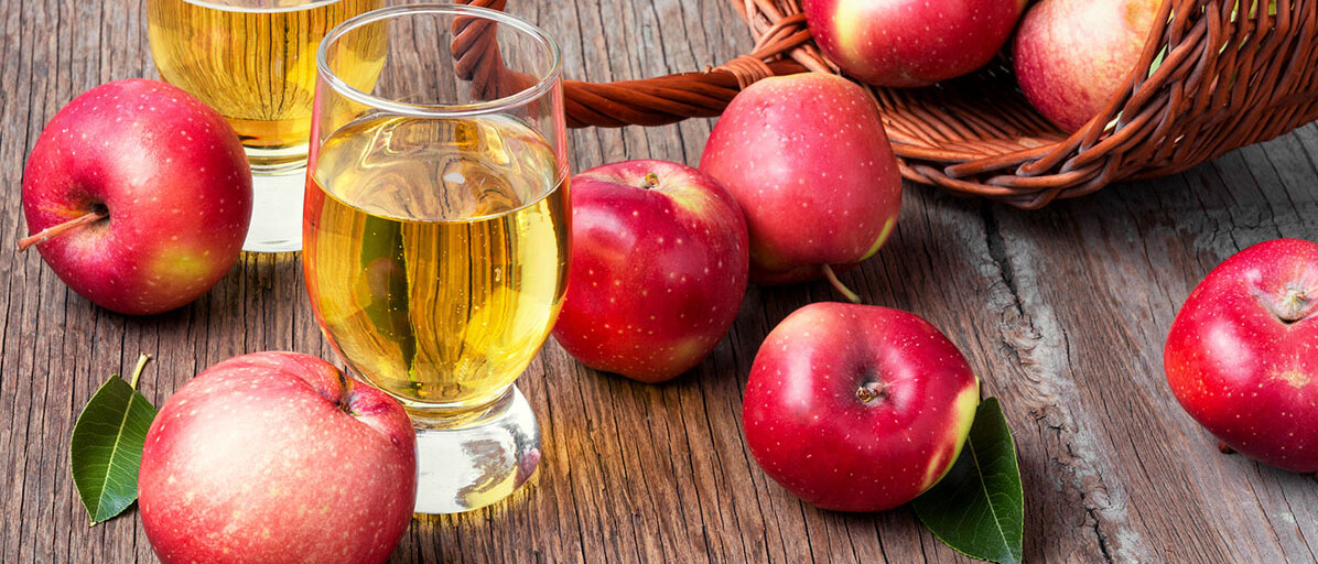 Apfelwein in einem Glas und drumerhum liegen rote Äpfel und ein umgefallener Holzkorb aus dem Äpfel herausfallen