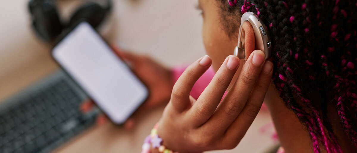 Junge Schwarze Frau mit pinken Rasta-Zöpfen trägt ein goldenes Hörgerät hinter dem Ohr und blickt auf ihr Handy. 