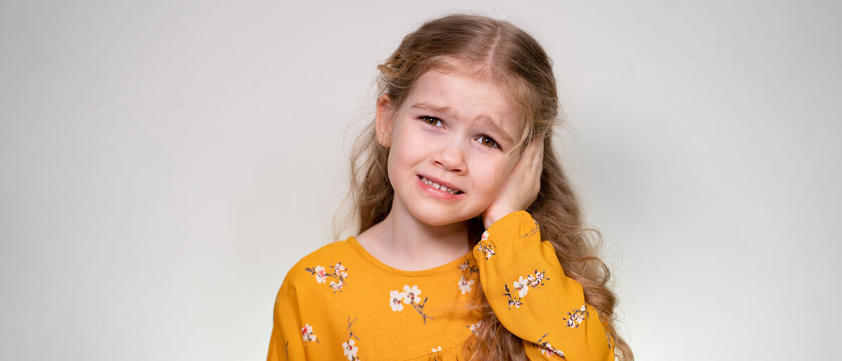 Ein kleines Mädchen hält sich die Hand ans Ohr und verzieht das Gesicht weinerlich.