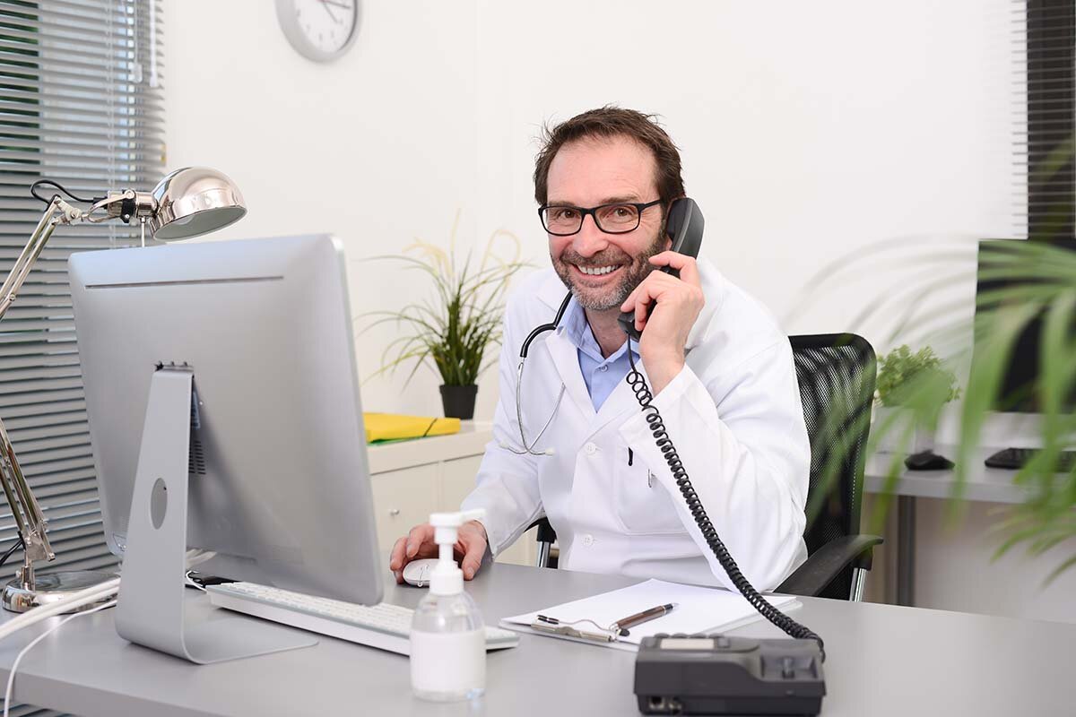 Ein Arzt im Kittel und mit Brille sitzt am Schreibtisch, vor ihm ein Computermonitor. In der linken Hand hält er einen schwarzen Telefonhörer. Er lächelt in die Kamera. 