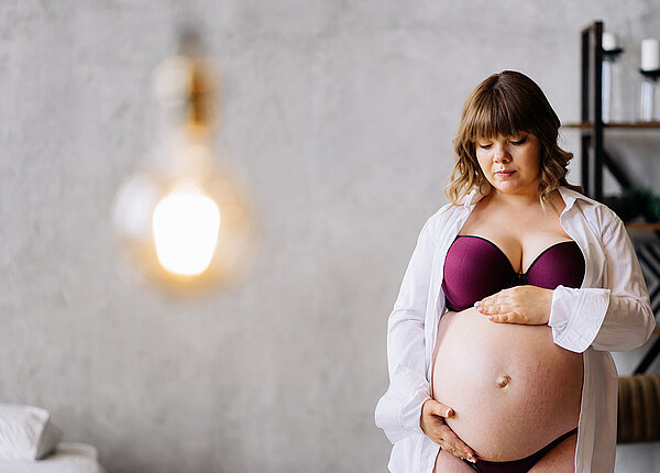 Eine schwangere Frau in Unterwäsche und offenem Hemd streichelt ihren Bauch.